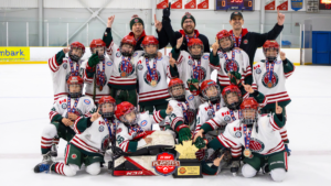Youth hockey team in white jerseys with red and green trim celebrate on the ice, medals around necks, two coaches raised arms behind them, trophy displayed front and center.