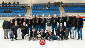 Hockey team posing on the ice with medals and a trophy after a playoff win, blue seats in the arena backdrop.