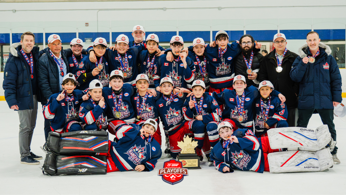 Youth hockey team poses on the ice after winning playoffs, wearing medals and caps with a trophy nearby.