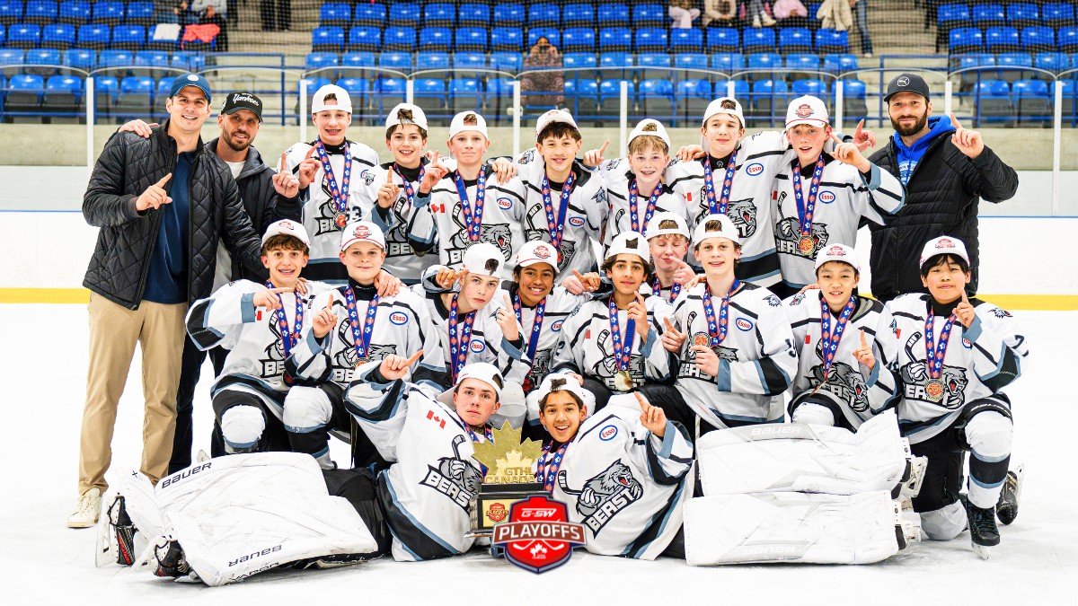 Youth hockey team posing on the rink with medals and a trophy, flanked by two coaches, blue arena seats in the background.